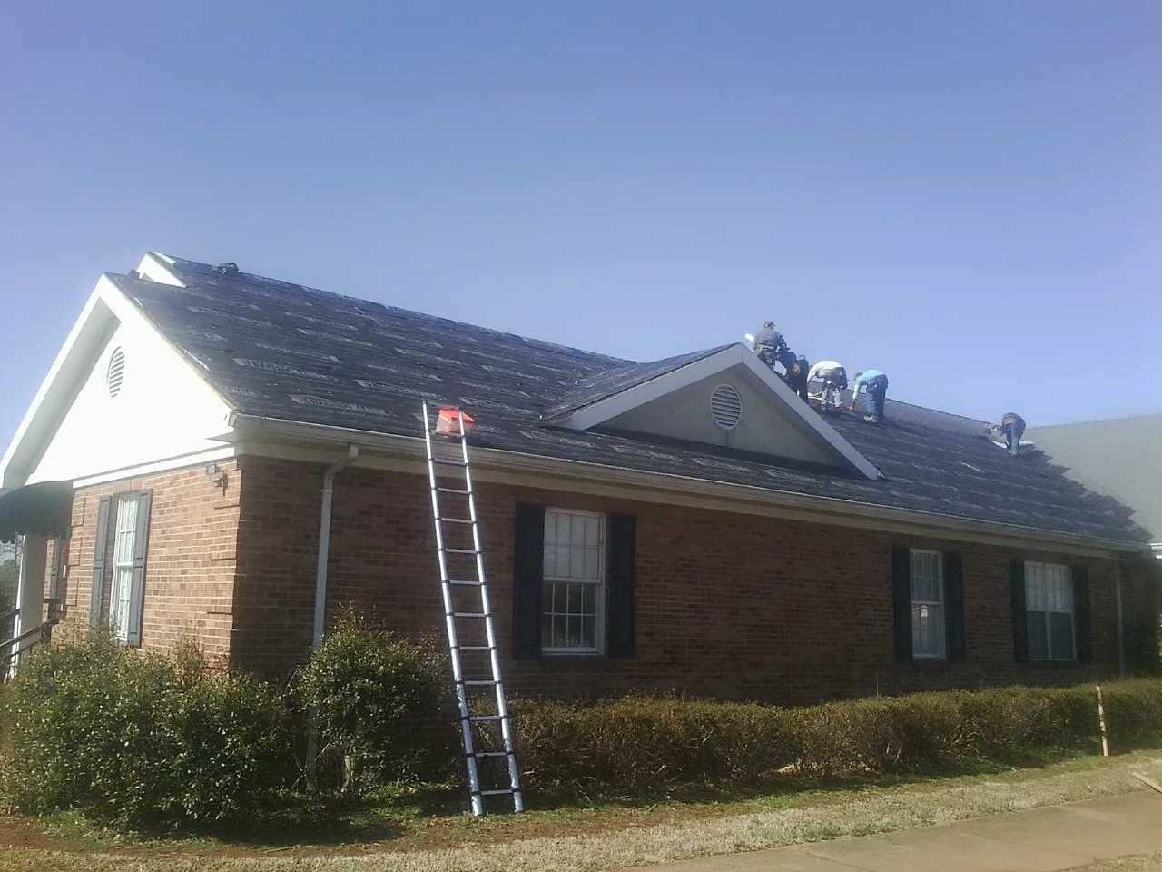 Crew tearing shingles off a brick ranch roof with a ladder set — Metal Roof Repair work by our crew in Lester