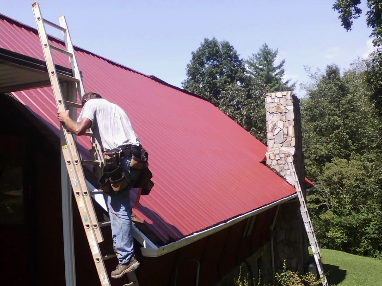 Roofer on a ladder beside a red metal roof and stone chimney — Soffit & Fascia Repair by Roofing Brothers in Lester